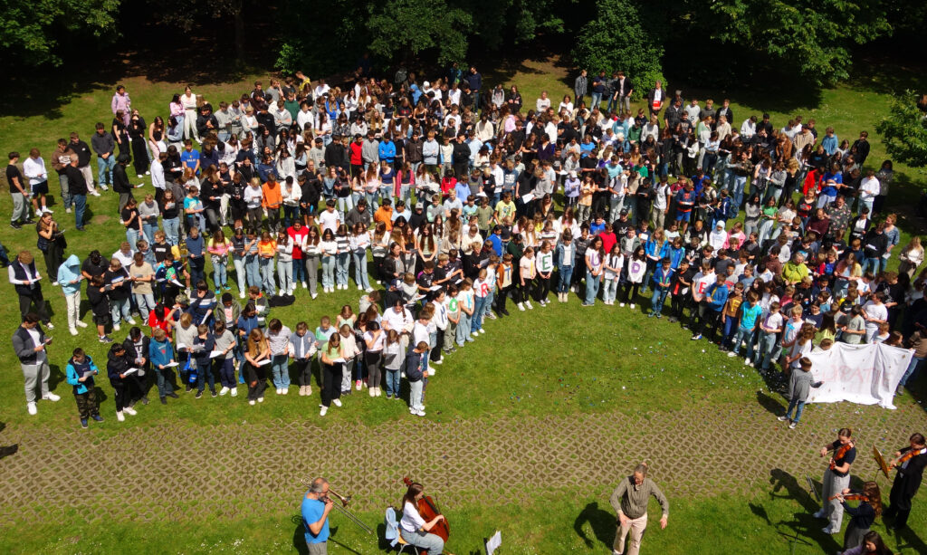 Ein Foto aus der Vogelperspektive, wie die gesamte Schüler-und Lehrerschaft auf dem Schulhof versammelt ist.