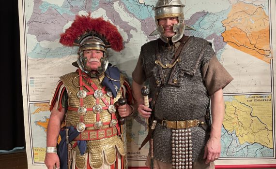 Die beiden Darsteller verkleidet als Legionäre vor einem Plakat zum römischen Reich